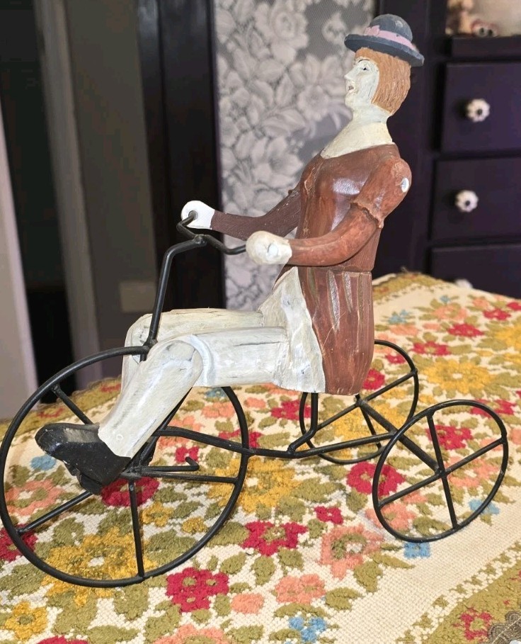 Vintage-style hand-carved folk art figurine of a woman on a tricycle.